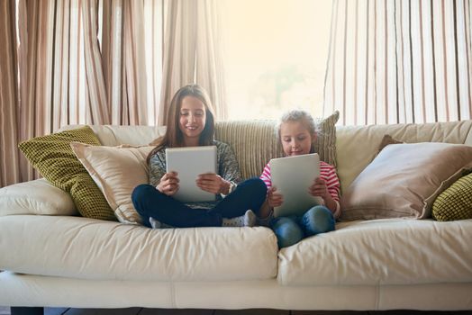 Technology is a great babysitter. two little girls playing with their tablets while sitting on a sofa at home.