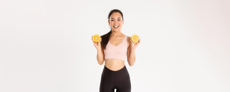 Sport, wellbeing and active lifestyle concept. Portrait of smiling healthy and slim asian girl advice eating healthy food for breakfast, gain energy for workout, hold two halves of orange