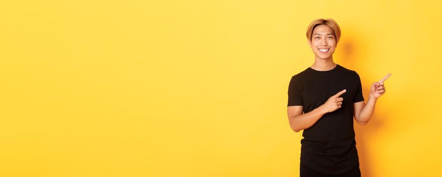 Portrait of satisfied smiling blond guy, asian student pointing fingers right, showing way, standing yellow background