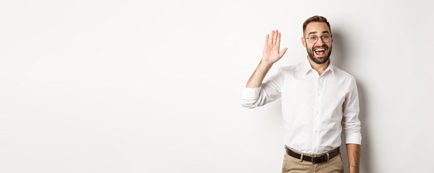 Friendly smiling man in glasses saying hello, waving hand in greeting, standing over white background