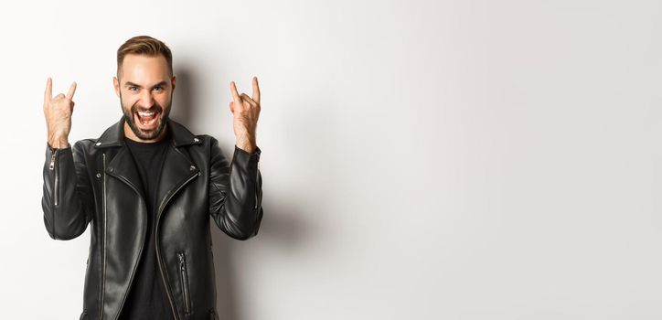 Cool adult man in black leather jacket, showing rock on gesture and tongue, enjoying music festival, standing over white background