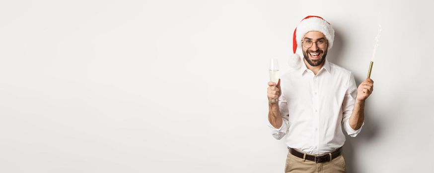 Winter holidays and celebration. Handsome bearded man having New Year party, holding firework sparkle and champagne, wearing santa hat, white background