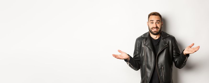 Confused bearded man in black leather jacket asking for advice, looking clueless and shrugging, standing over white background