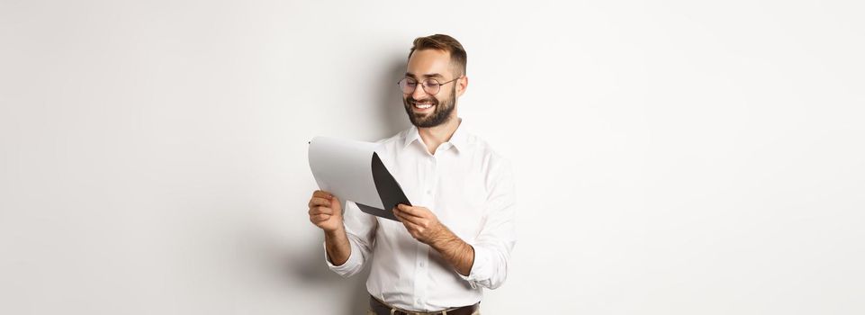 Businessman looking satisfied at documents, reading report and smiling, standing over white background