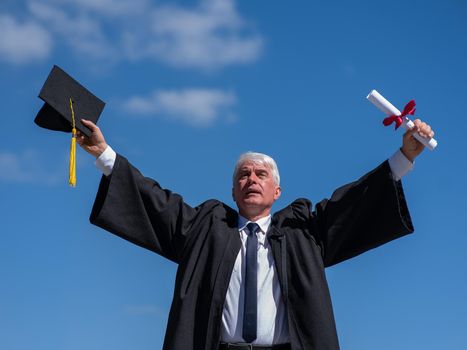 Elderly male graduate rejoices in receiving a diploma against a blue sky.