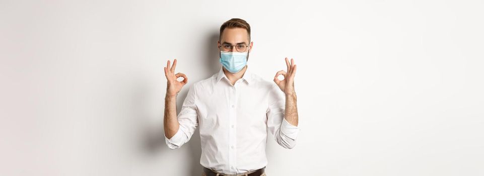 Covid-19, social distancing and quarantine concept. Confident businessman wearing medical mask and showing okay signs in approval, white background