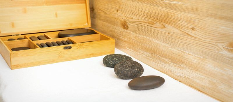 Black massage stones lying near the wooden box with massage rocks on the towel on the table.
