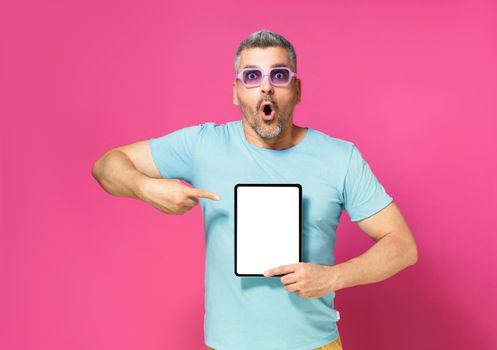 Great offer pointing at digital tablet white screen handsome man looking at camera with shock facial expression wearing casual blue shirt and sunglasses isolated on pink background. App advertisement