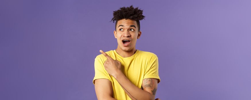 Portrait of impressed hispanic guy seeing something interesting, pointing finger and looking upper left corner, discuss strange thing upwards, standing purple background curious