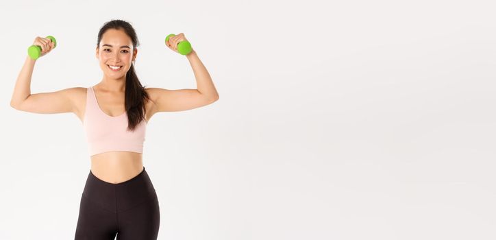 Fitness, healthy lifestyle and wellbeing concept. Portrait of strong and happy female athlete, asian girl workout at home during coronavirus, lifting dumbbells to gain muscles, white background
