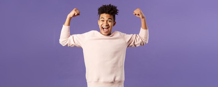 Portrait of young happy man got scholarship, applied to cool university, raise hands up flex biceps like champion, triumphing from great news, achieve goal and rejoicing, purple background