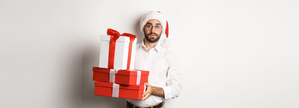 Merry christmas, holidays concept. Confused man in Santa hat holding pile of presents, found gifts under xmas tree, standing against white background