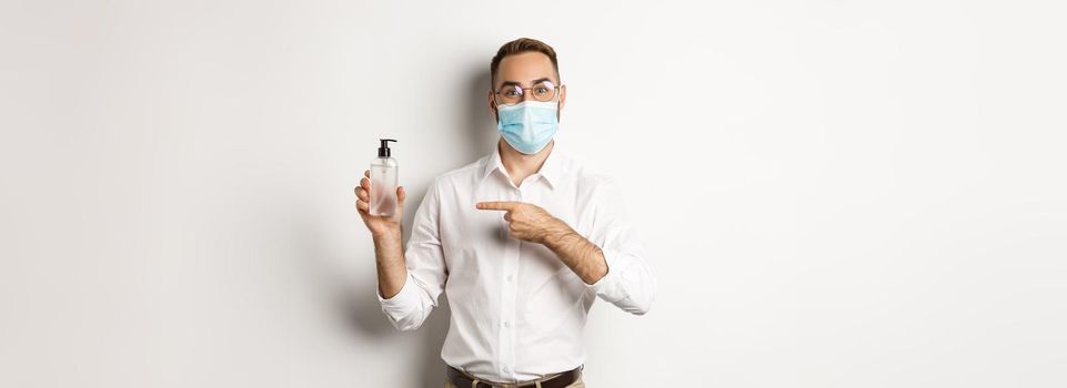 Covid-19, social distancing and quarantine concept. Office worker in medical mask pointing at hand sanitizer, showing antiseptic, white background
