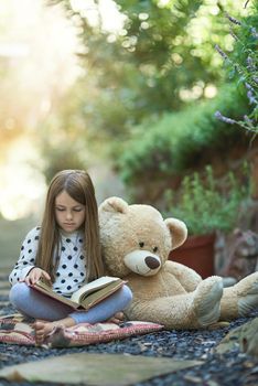 Lost in the wonder of her favourite story. a little girl reading a book with her teddy bear beside her.
