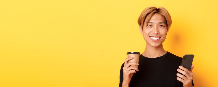 Close-up of handsome young asian guy smiling happy, using smartphone and drinking takeaway coffee, standing over yellow background