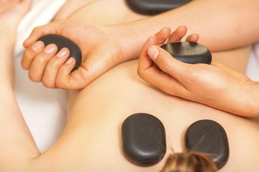 Hot stone massage on the female back with hands of masseur holding black massage stones in spa salon.
