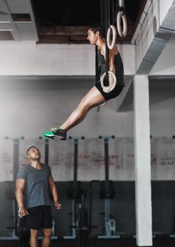 Motivated by her trainer. Full length shot of a young woman working out on the gymnastics rings while her trainer looks on.