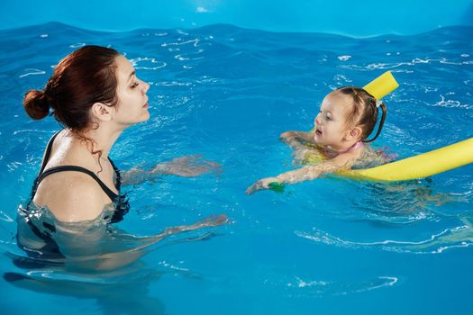 Preschool girllearning to swim in pool with foam noodle with young trainer