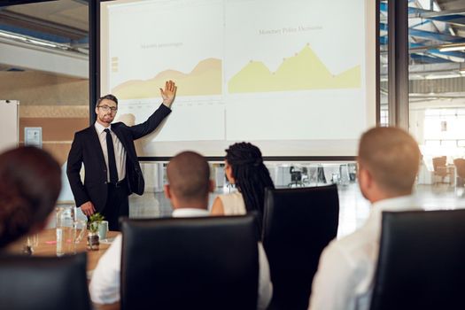 Explaining the figures clearly and concisely. an executive giving a presentation on a projection screen to a group of colleagues in a boardroom.