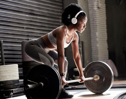 Power, fitness and headphones of a woman with barbell weight lift exercise, workout or training in wellness gym. Sports black woman listening to motivation music for muscle, strength and health