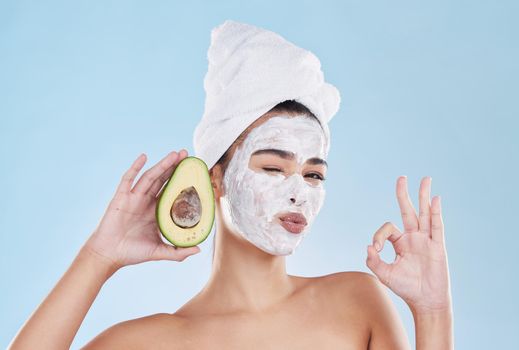 Face mask, avocado and woman with a ok sign with health, wellness and organic lifestyle in studio. Girl doing fresh, clean and natural selfcare routine while holding fruit for nutrition and diet.