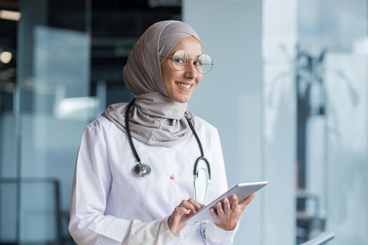 Portrait of a Muslim female doctor in a hijab with a tablet in her hands, the doctor works in the office of a modern clinic, smiles and looks at the camera