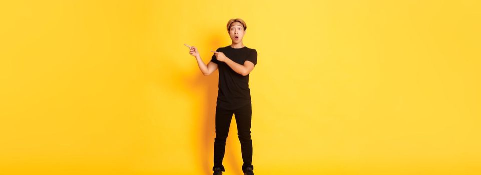 Full-length of amazed and impressed attractive asian guy pointing fingers upper right corner, showing logo, yellow background
