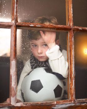 Cant it rain some other time. a sad little boy holding a soccer ball while watching the rain through a window.