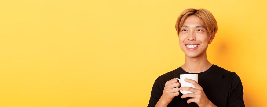 Close-up of happy smiling handsome blond asian guy, looking away with dreamy nostalgic look while drinking coffee, standing over yellow background