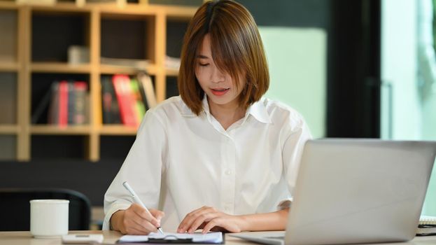 Young creative woman working in contemporary office, using laptop and making notes on notebook