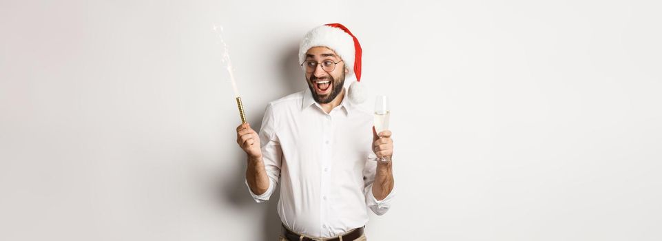 Winter holidays and celebration. Excited man celebrating New Year eve with fireworks sparkles and drinking champagne, wearing santa hat, white background