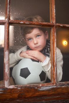 Why does it always rain when I want to play. a sad little boy holding a soccer ball while watching the rain through a window.
