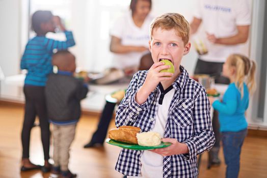 Nutrition above all. Portrait of a little boy with a plate of food with volunteers serving food in the background.