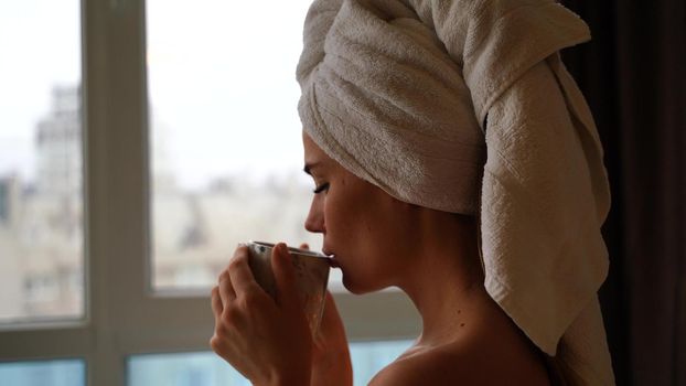 Middle aged woman looks good with bare shoulders in a white towel on her head holds a cup and drinks coffee or tea against the window
