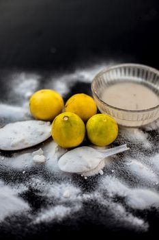 Baking soda face mask in a glass bowl on wooden surface along with some baking soda sprinkled on the surface and lemons also on surface. Used to blemishes skin instantly.