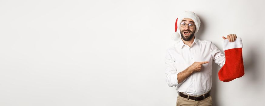 Merry christmas, holidays concept. Happy adult man receive gifts in xmas sock, looking excited, wearing santa hat, white background
