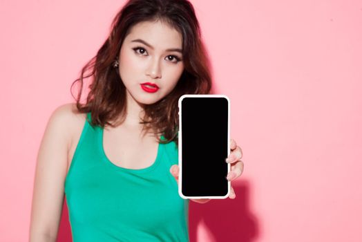 Cheerful cute young woman holding and pointing on blank screen mobile phone over pink background