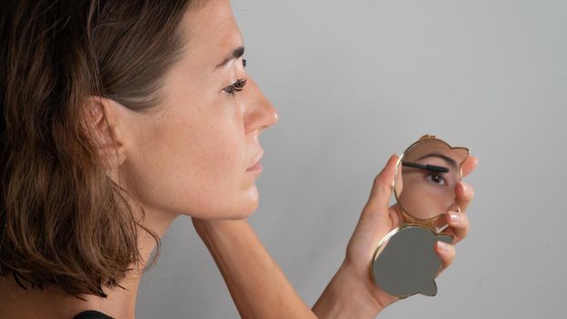 A woman dyes her eyelashes with mascara in a small mirror, a reflection