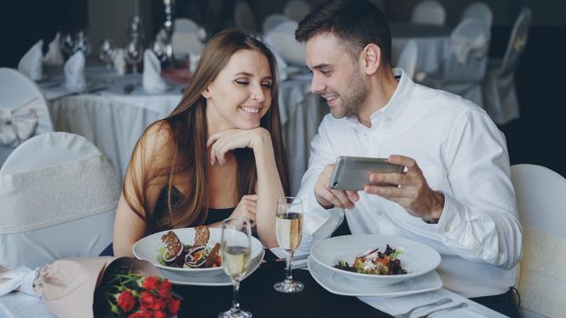 Happy attractive young people are watching smartphone together, smiling and talking while having romantic dinner in restaurant. Modern technologies and romance concept.