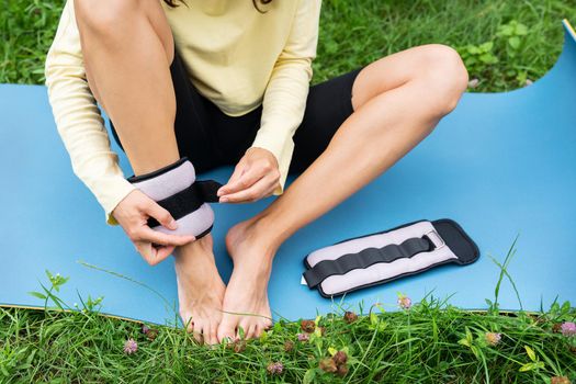 A sports girl does a stretch in nature in the fresh air, the girl hangs weights on herself for a better study of the exercises. A woman is doing yoga in the park, sitting on the grass on a yoga mat