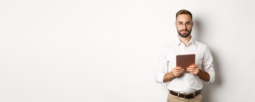 Serious employer working with digital tablet, reading in glasses, standing over white background
