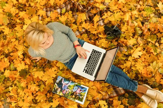 Cute woman with laptop in the autumn park. Beauty nature scene with colorful foliage background, yellow trees and leaves at fall season. Autumn outdoor lifestyle. Happy smiling woman on fall leaves