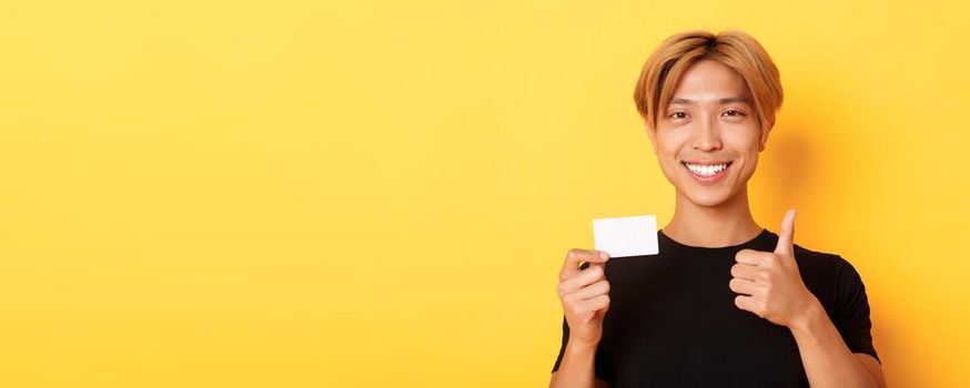 Close-up of satisfied asian guy recommend bank, showing credit card and standing over yellow background