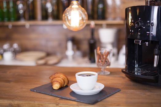 Coffee served with croissant next to black coffee machine on the bar counter