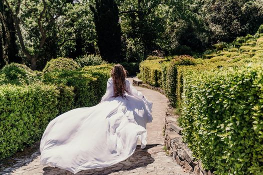 a beautiful woman with long brown hair and long white dress runs along a path along beautiful bushes in the park rear view