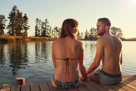 Enjoying the last rays of a summers day. an affectionate young couple in swimsuits sitting on a dock at sunset.