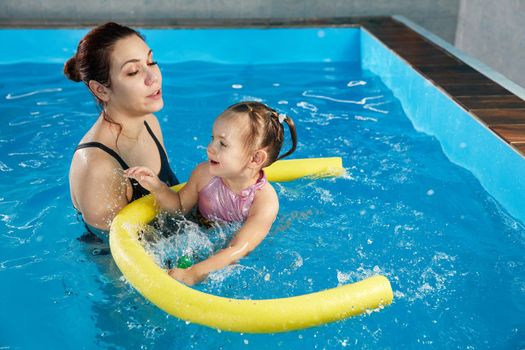Preschool girllearning to swim in pool with foam noodle with young trainer