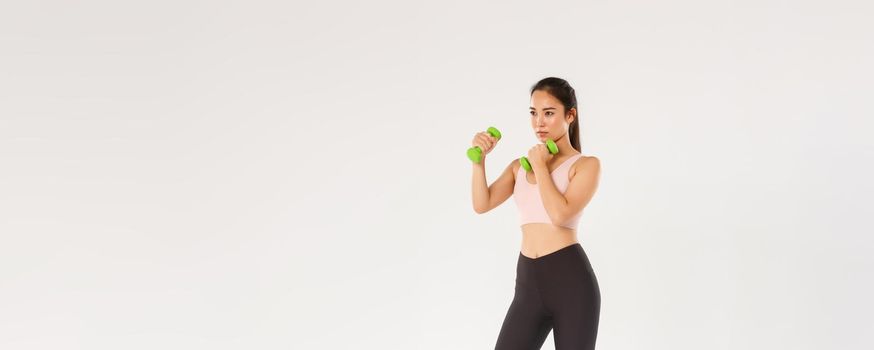 Sport, gym and healthy body concept. Full length of serious-looking asian brunette sportswoman, female athlete in sports bra and leggings lifting dumbbells, fitness exercises over white background
