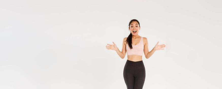 Full length of surprised and amazed female athelte, asian fitness girl in sportsbra and leggings looking wondered, smiling excited and spread hands sideways, find out about gym special discounts