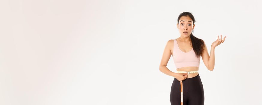 Fitness, healthy lifestyle and wellbeing concept. Surprised asian girl on diet, sportswoman wrap measuring tape around waist and look impressed as lose weight with workout